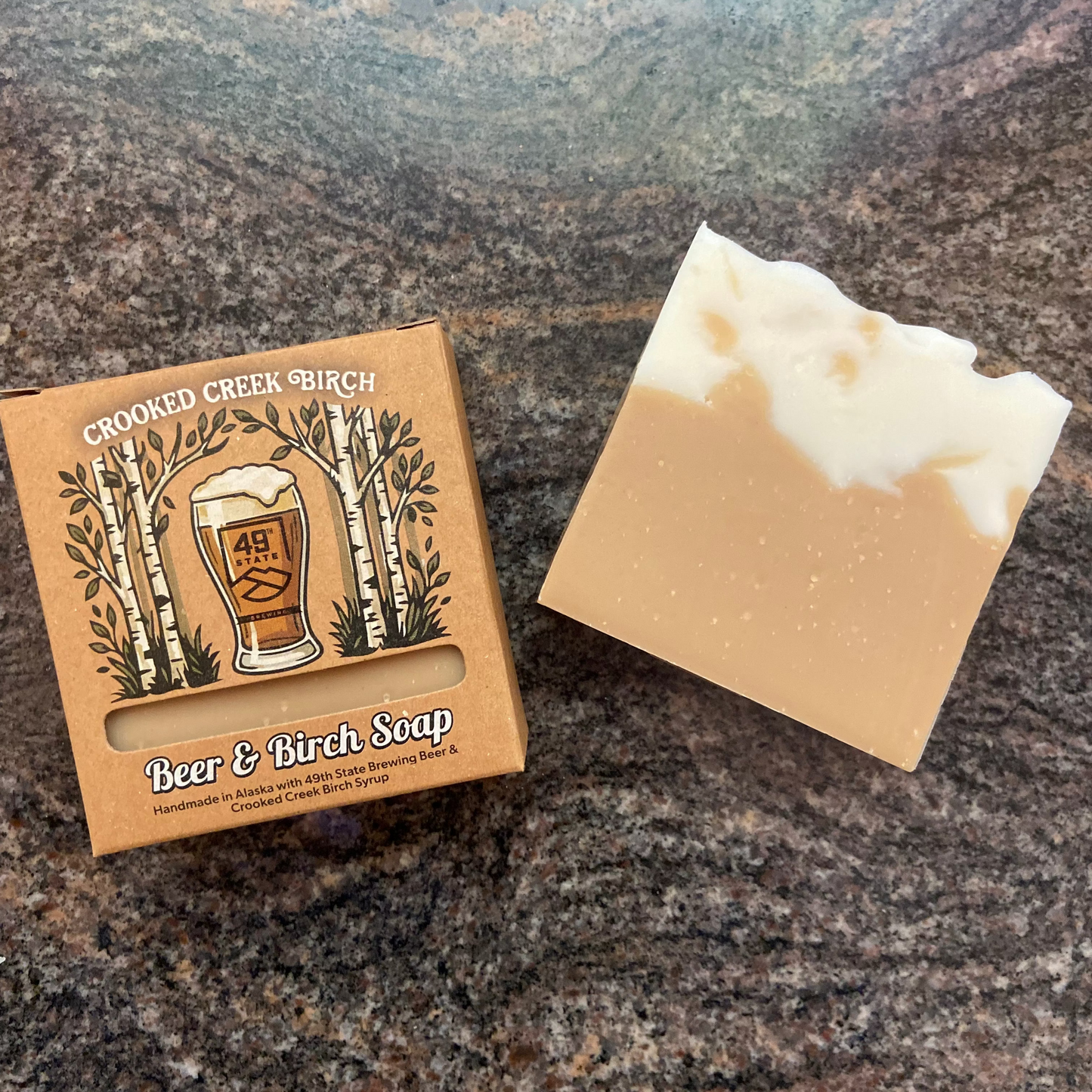 A bar of Crooked Creek Birch Beer & Birch Soap next to its brown cardboard packaging box, displayed on a dark, textured granite surface.